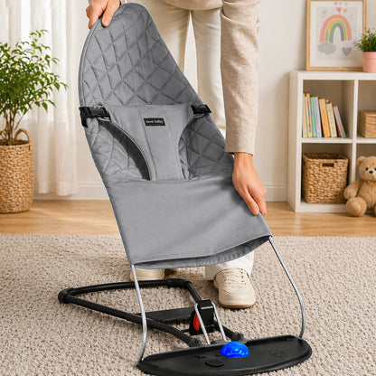 A person is holding a gray quilted baby bouncer chair with a black metal base and footrest in a bright, modern nursery. The brand name 'Bebamour' is visible on a small black label on the front of the fabric.