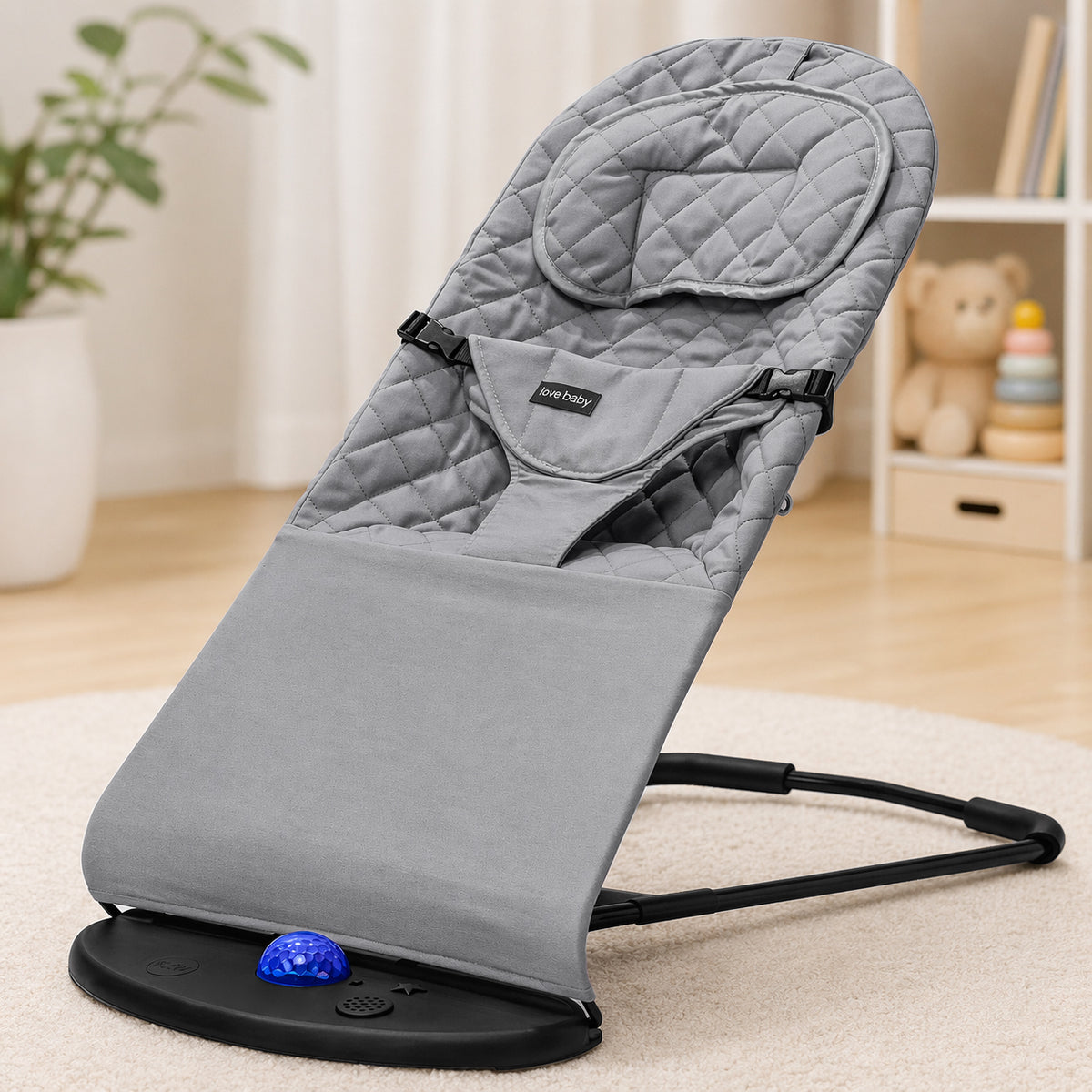 A gray padded baby bouncer chair with a black support base and harness straps, photographed in a softly lit nursery setting on a light rug.
